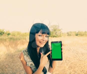 Brunette girl holds a tablet touch pad computer gadget with green screen outside in the summer and point at screen