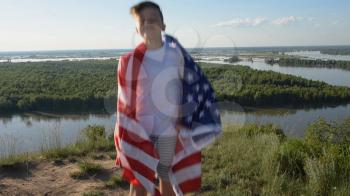 Blonde boy waving national USA flag outdoors over blue sky at the river bank.