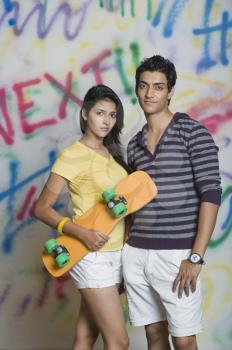 Portrait of a couple standing with a skateboard in front of a graffiti covered wall