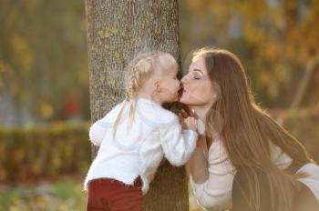 A young mother in a sweater and her cute daughter are having fun on a walk in the autumn Park. Mom plays with her daughter next to a tree and kisses her