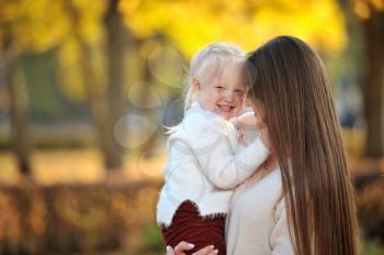 A young mother in a sweater holds her cute blonde daughter in her arms during a walk in the autumn Park, against the background of fallen leaves and trees