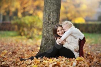 A young mother in a sweater and her cute daughter are having fun on a walk in the autumn Park. Mom sits on fallen leaves and her cute blonde daughter hugs her