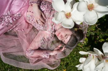 A young girl in a lush pink dress lies with her eyes closed under a magnolia tree with large white flowers. Girl, dreamy image and blooming magnolia