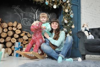 Young mother in jeans and her little daughter on a red rocking horse sit against the background of the Christmas interior and smile