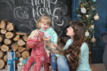 Young mother in jeans and her little daughter on a red rocking horse sit against the background of the Christmas interior and smile