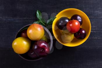 Colorful plums fruit in pot and plate on the dark background top view