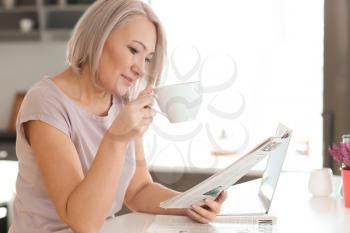 Mature woman drinking coffee while reading newspaper at home�