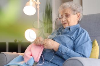 Senior woman sitting on armchair while knitting sweater at home�