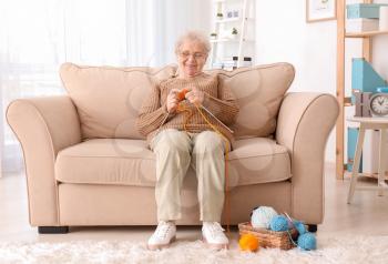 Senior woman sitting on sofa while knitting sweater at home�