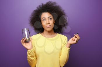 Young African-American woman holding glass jar with coins on color background. Savings concept�