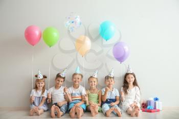 Cute little children in Birthday hats and with balloons sitting on floor near light wall�