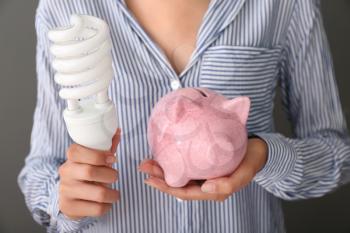 Woman holding piggy bank with light bulb, closeup. Electricity saving concept�
