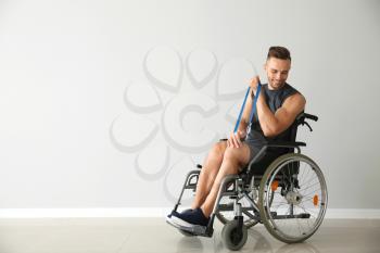 Sporty man training with elastic band while sitting in wheelchair against light wall�