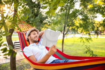 Handsome young man with book resting in hammock outdoors�