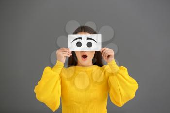 Emotional young woman hiding face behind sheet of paper with drawn eyes on grey background�