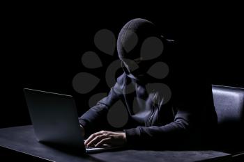 Professional hacker with laptop sitting at table on dark background�
