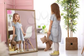 Cute little girl with teddy bear looking in mirror at home�