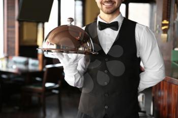 Young male waiter with tray and cloche in restaurant�