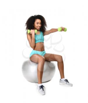Sporty African-American woman training with fitball and dumbbells on white background�