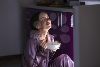 Happy young woman eating food near refrigerator at night�