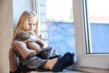 Homeless little girl with teddy bear sitting on window sill�