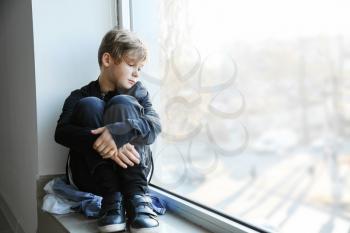 Homeless little boy sitting on window sill indoors�