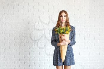 Beautiful young woman with bouquet of mimosa flowers against white brick wall�
