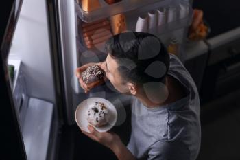 Young man eating food near refrigerator at night�