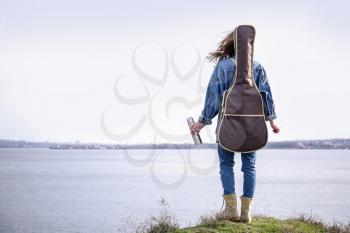 Woman with thermos and guitar near river, back view�