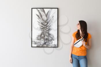 Beautiful young woman standing near picture hanging on light wall�