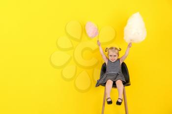 Cute little girl with cotton candy sitting on chair against color background�