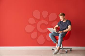 Handsome young man with laptop sitting on chair near color wall�