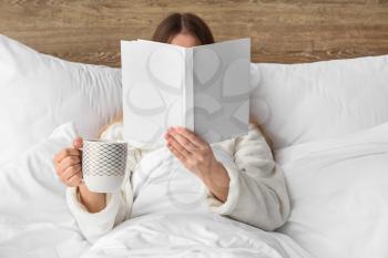 Young woman with cup of hot beverage reading book in bed�