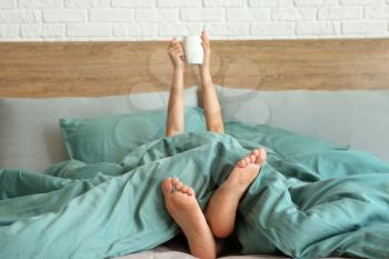 Young woman with cup of hot beverage lying in bed�
