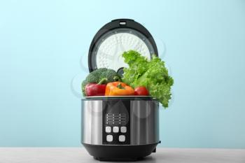 Modern multi cooker with vegetables on table against color background�