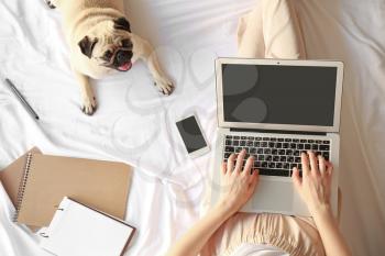 Young woman with laptop and cute pug dog resting on bed�