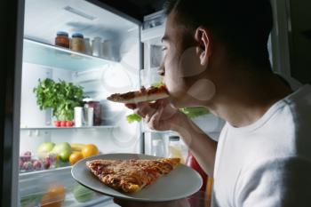 Young man eating pizza near refrigerator at night�