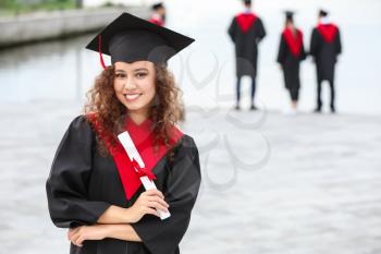 Happy African-American student in bachelor robe and with diploma outdoors�
