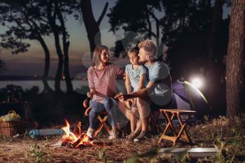 Family roasting sausages over campfire in evening�