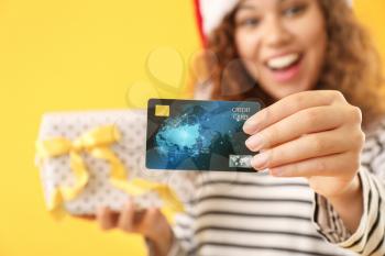 Happy African-American woman in Santa hat, with gift and credit card on color background�