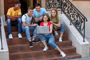 Portrait of young students with laptop sitting on stairs outdoors�