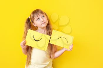 Thoughtful little girl with drawn smiling and upset mouths on sheets of paper against color background�