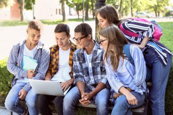 Young students with laptop sitting on bench outdoors�
