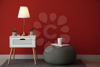 Modern table with lamp and pouf near color wall in room�