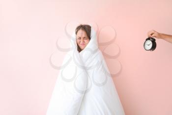 Displeased young woman wrapped in blanket and male hand with alarm clock on color background�