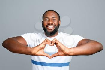 Happy African-American man showing heart shape with hands on grey background�