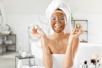 Beautiful young woman with facial mask and cucumber slices in bathroom�