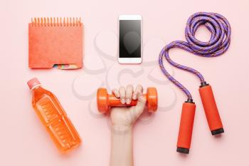 Female hand with dumbbell, bottle of water, mobile phone, notebook and jumping rope on color background�