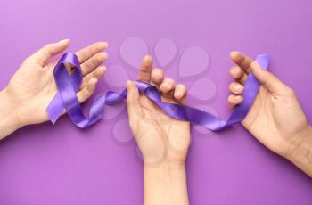 Female hands with purple ribbon as symbol of World Cancer Day on color background�