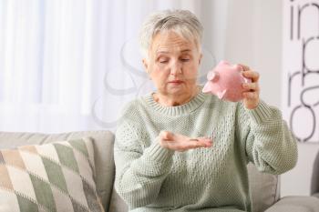 Sad senior woman with empty piggy bank at home�